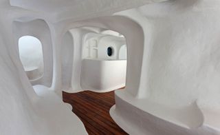 Interior view of The Original Dwelling by Atelier Van Lieshout - a cave-like structure with white walls and wood flooring