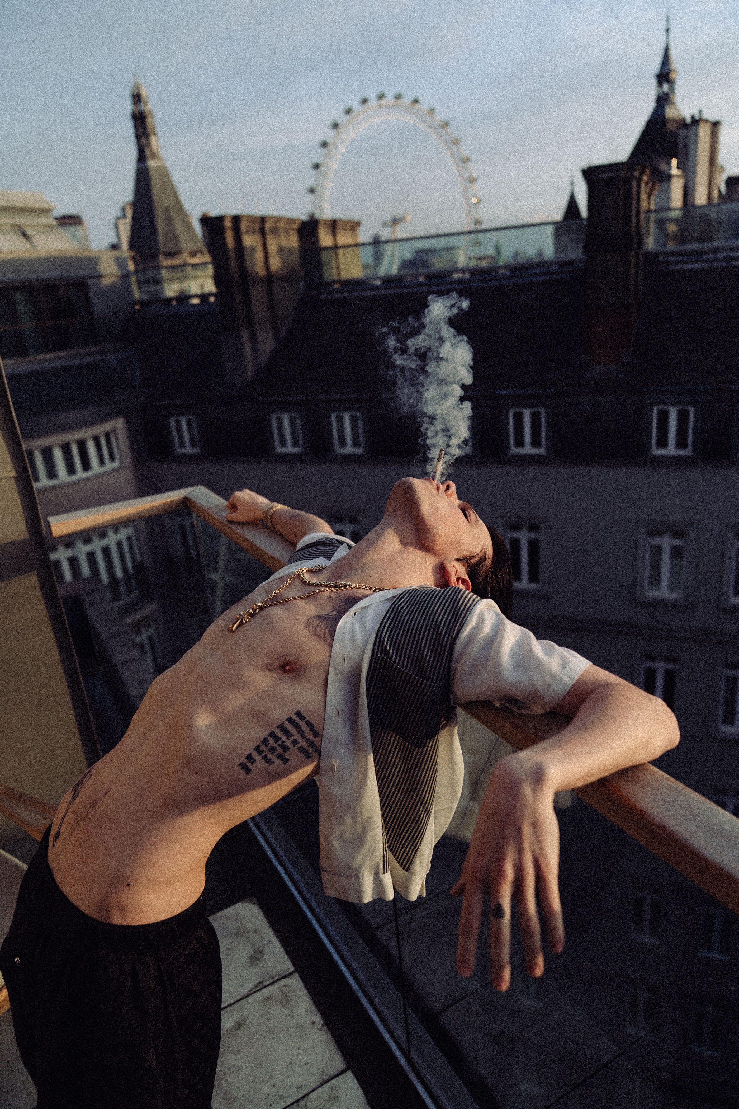 A portrait of Yungblud smoking leaning backwards on a balcony with the London Eye in the background