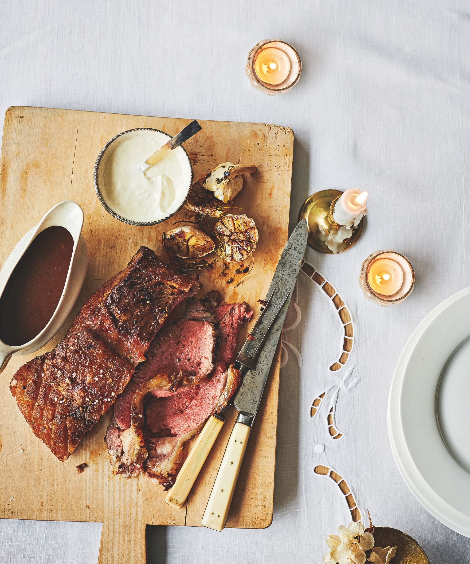 Roasted meat on a wooden chopping board on a white tablecloth, with a white gravy boat, carving knife and fork, candles, and a pile of white plates.