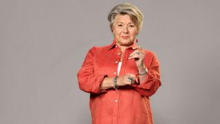 A posed shot of Anne Lloyd (Barbara Flynn) standing in front of a plain grey background. Anne is wearing a bright red shirt and holding her left arm up towards her shoulder, while her right arms reaches across her torso towards her left elbow.