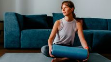 Woman sits cross-legged on yoga mat in front of sofa, holding a foam roller in both hands