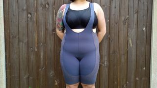 Some Rapha Women's Pro Team Powerweave Bib shorts worn over a black sports bra, modelled by a chubby tattooed woman, standing in front of a wooden fence. Due to their light colour and thin fabric, her curves are much more visible than with black shorts