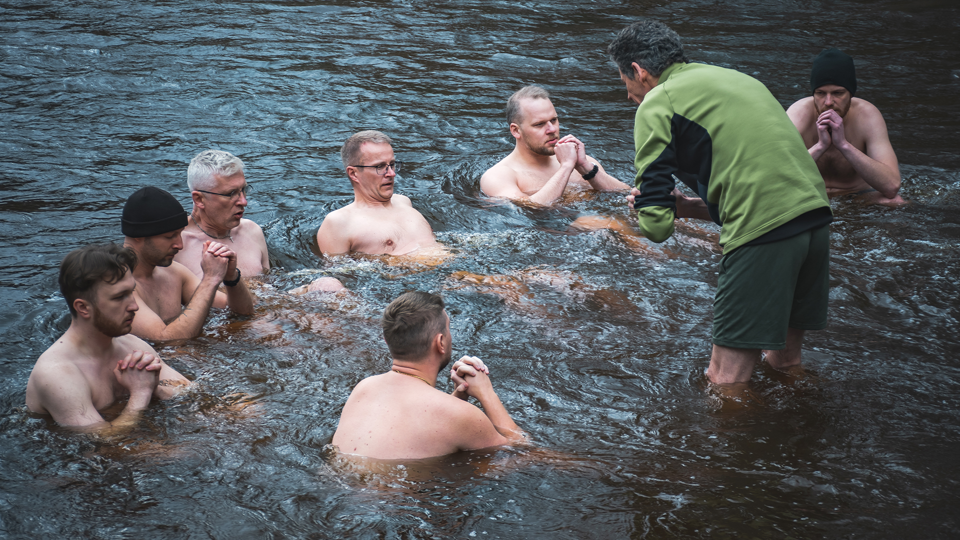 Group of men stand in a circle bathing a very cold river as an instructor in a green shirt stands with them