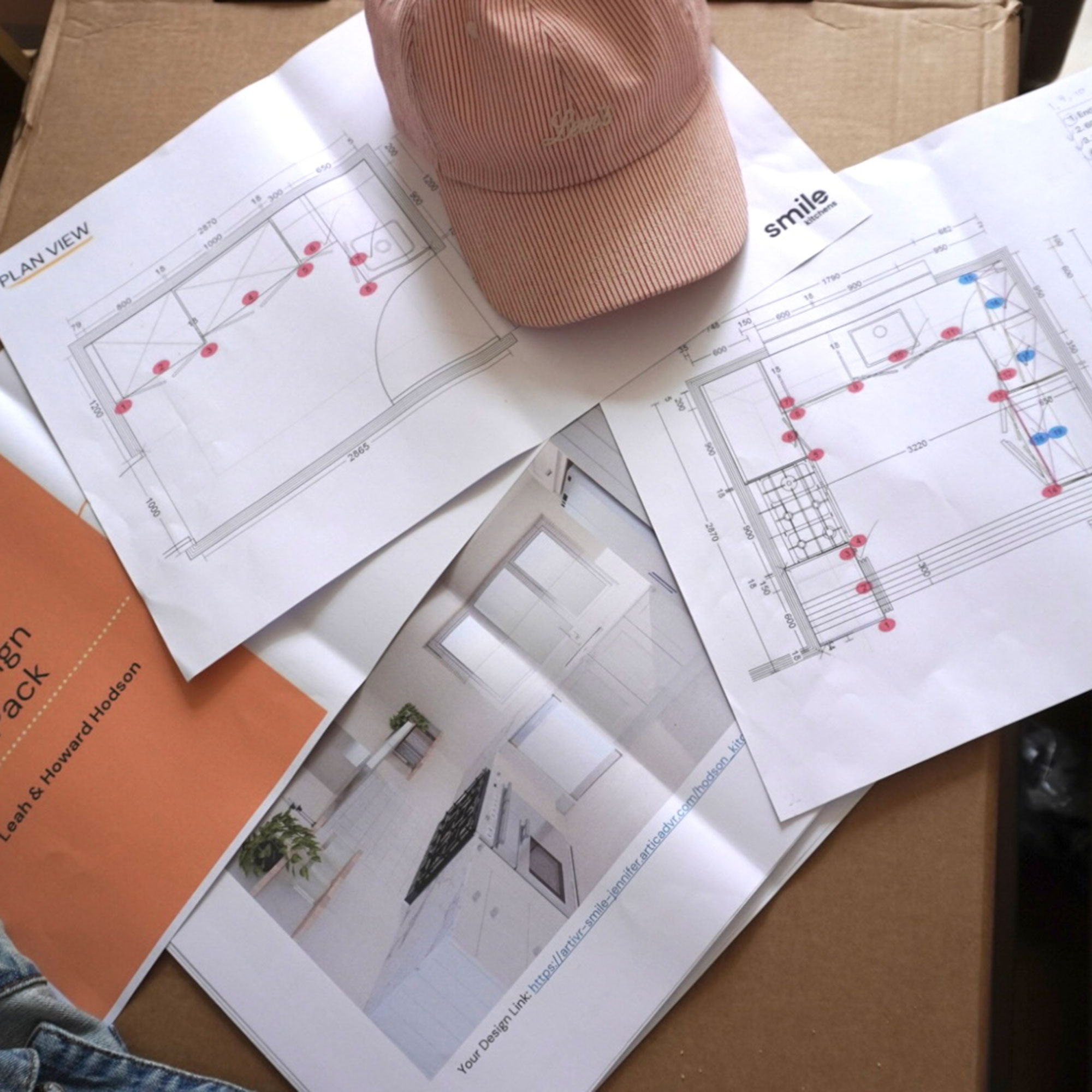 Paper plans for a kitchen laid out over a table, with a pink baseball cap resting on top