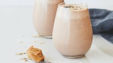 Two glasses filled with a beige smoothie next to a teaspoon of nut butter