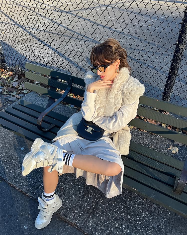 An image of @izzipoopi wearing Isabel Marant wedge trainers with a midi skirt, fur gilet and Celine triomphe bag.