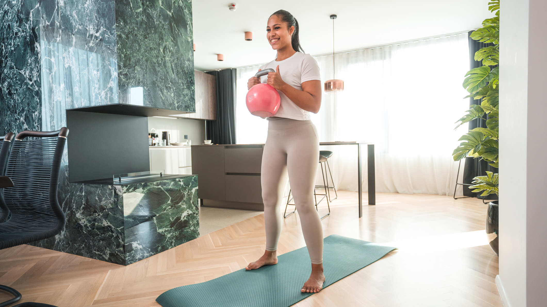 Woman stands in living area holding a kettlebell in front of her chest