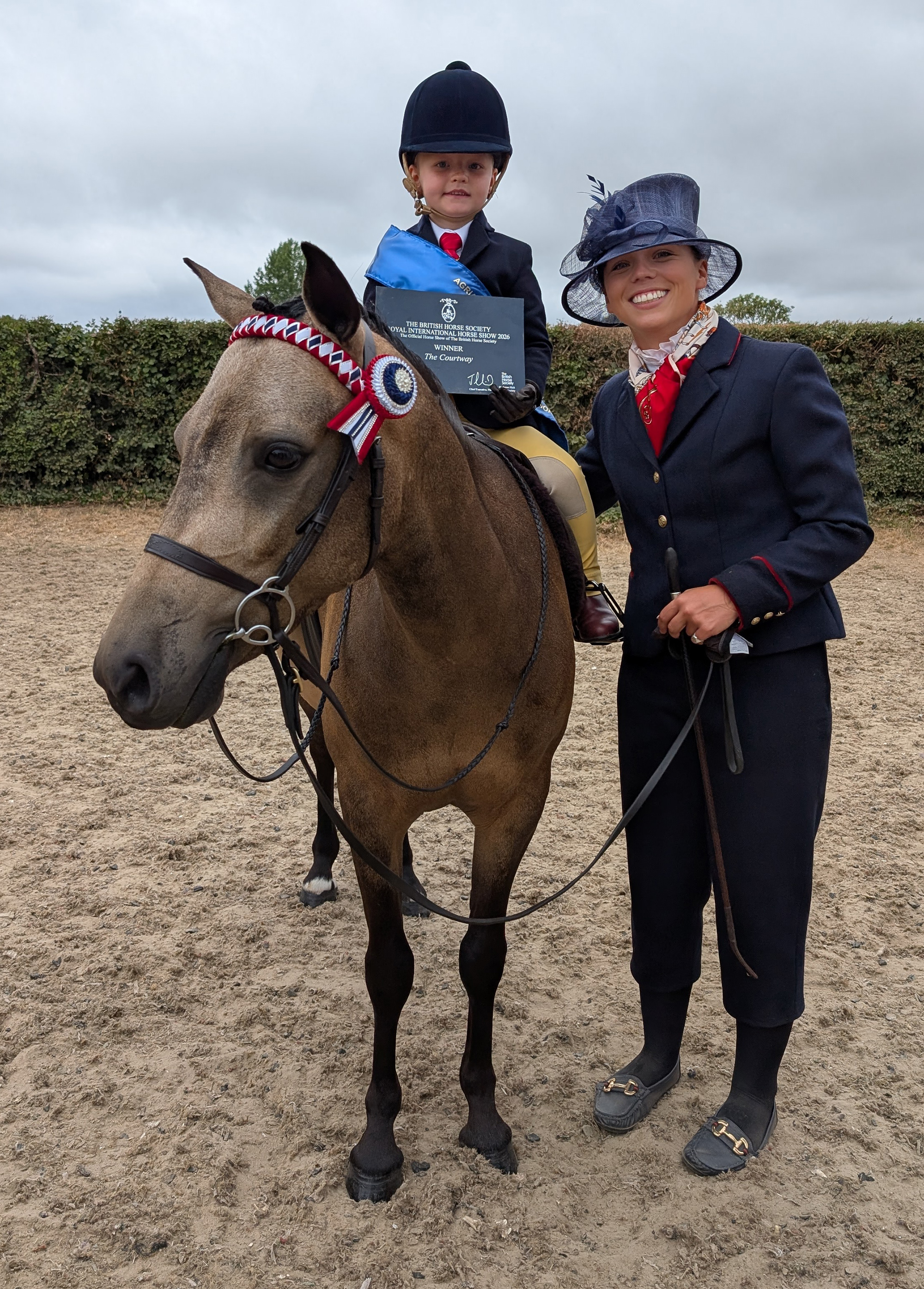 A bucksin lead rein show pony with tiny child rider wearing blue winner's sash and holding winner's card with handler dressed in navy blue standing alongside