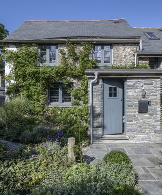 Small front garden ideas shown in front of a slate clad house with a blue door, planting and paving