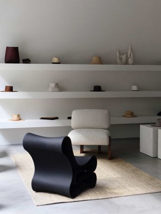 A minimalist shop features whimsically shaped furniture including black and white, 70s-inspired armchairs sitting on a rattan carpet, white, simple shelves complete with hats, and white podiums used as product displaying units.