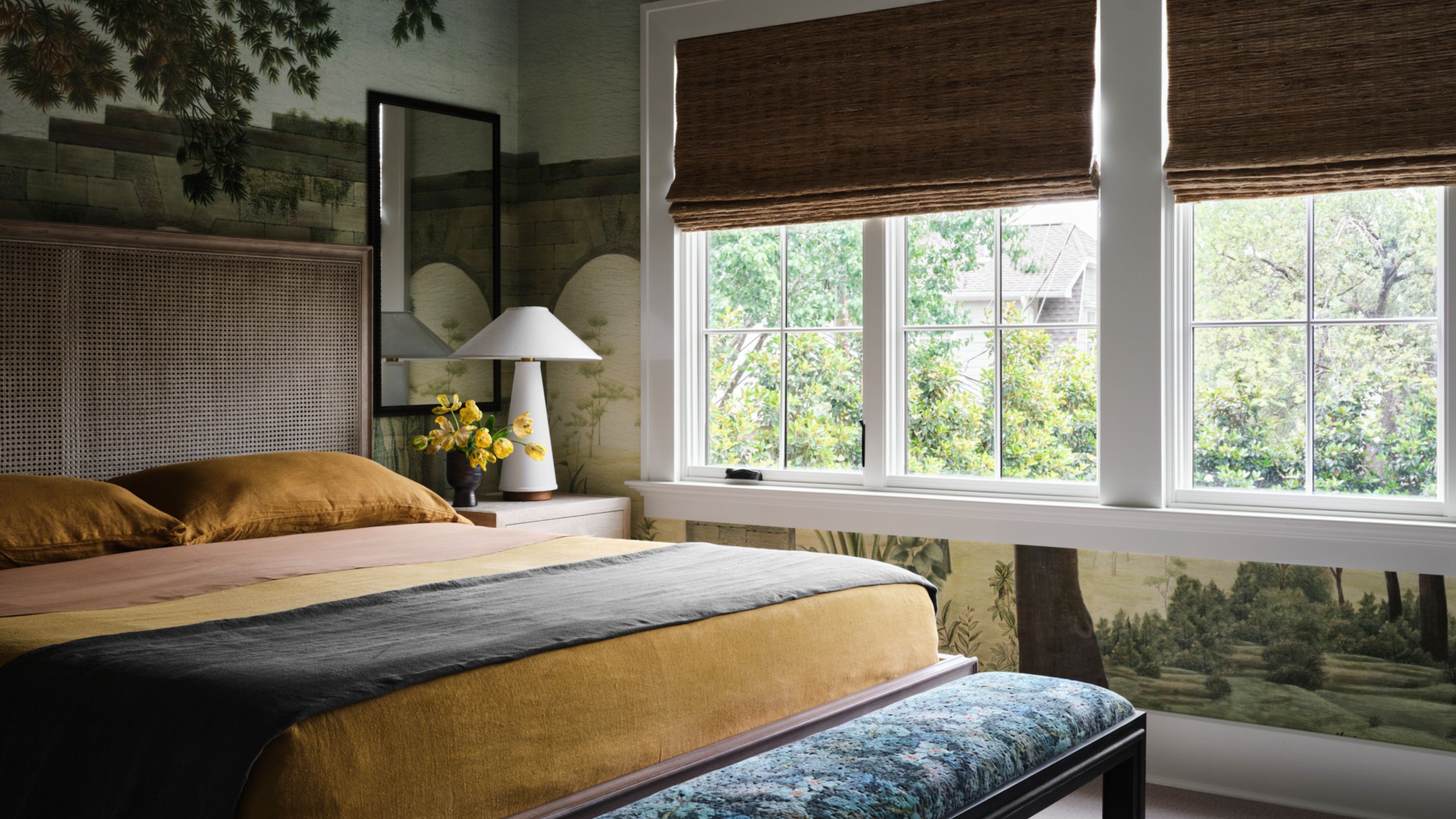 a bedroom with mural wallpaper, large bed with rattan bedhead, colorful sheets, a white lamp on the bedside table and a mirror, and woven bamboo blinds covering the windows