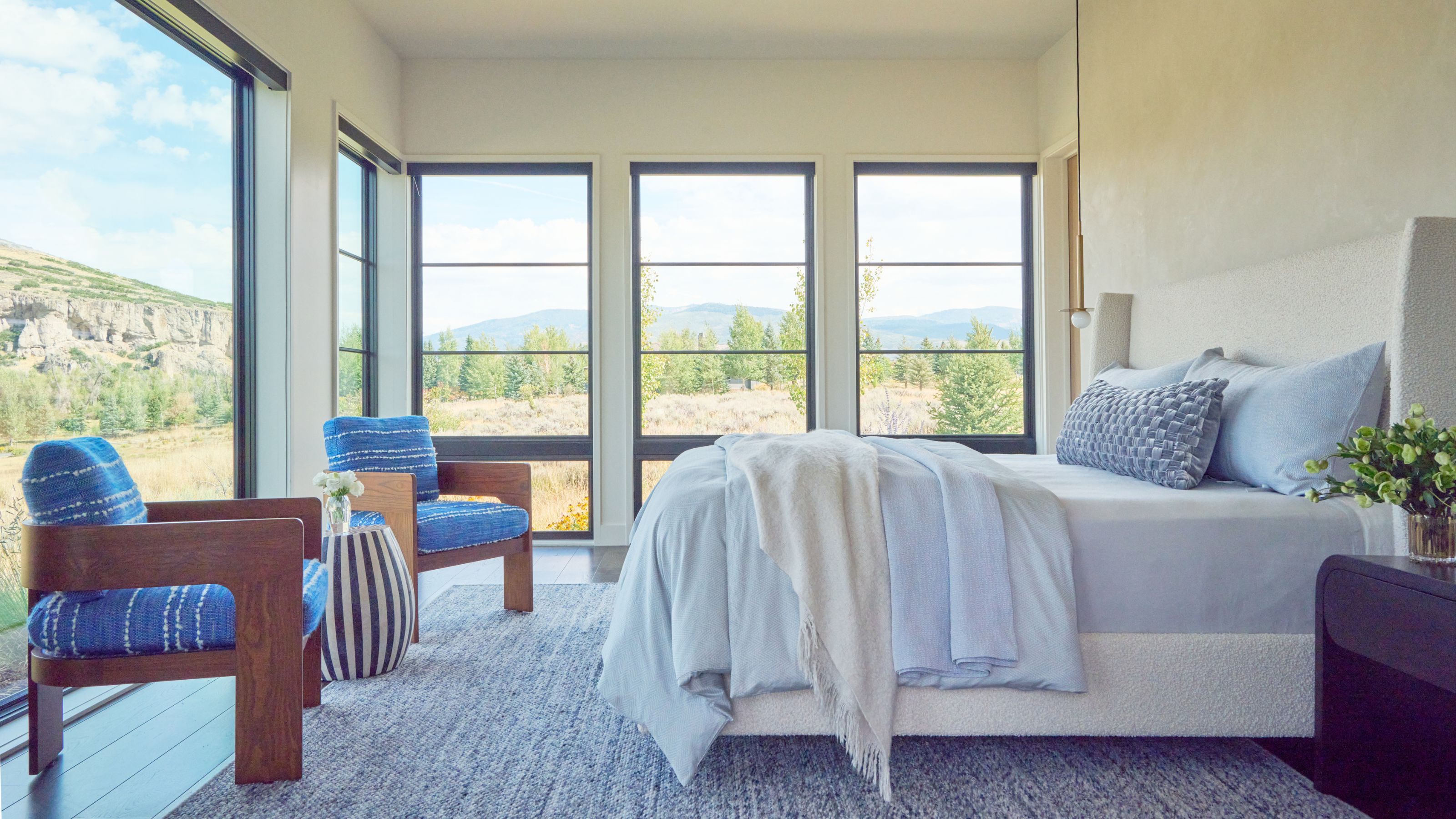 Light filled master bedroom that has large floor to ceiling windows and a large bed with cream headboard and white bedding. Two wooden chairs upholstered with blue cushions sit facing the bed