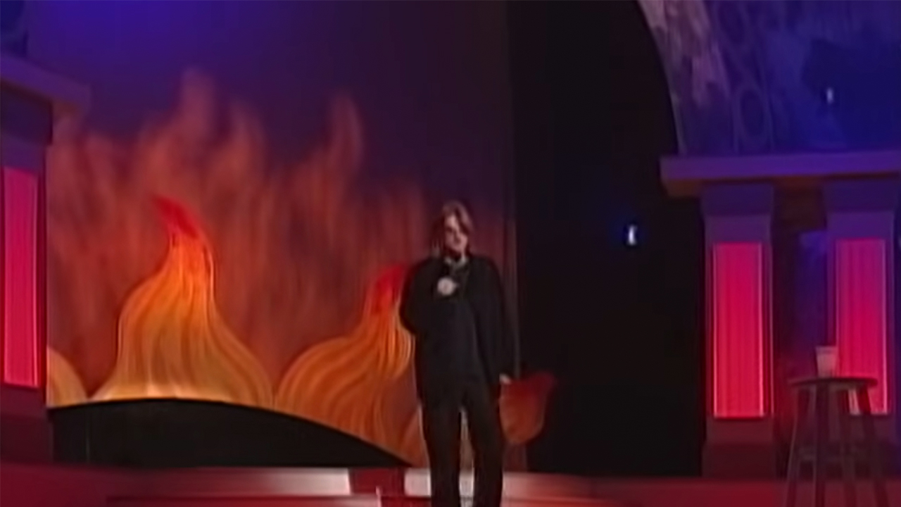 A long shot of Mitch Hedberg performing standup on a big stage