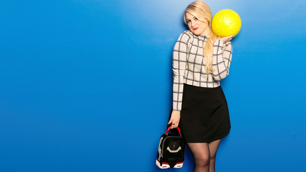 Meghan Trainor simles as she holds up a bright yellow bowling ball.