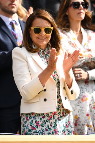 Carole Middleton wearing a white floral dress with a white blazer and yellow sunglasses smiling and clapping at Wimbledon