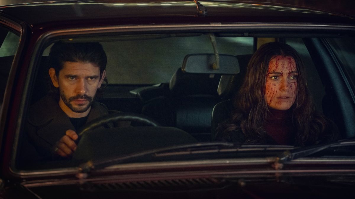 From left to right: Ben Whishaw looking serious and driving a car and Keira Knightley sitting in the passenger seat with blood on her face. 