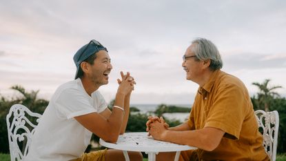 An adult son and his father sit at a table outside and laugh while talking.