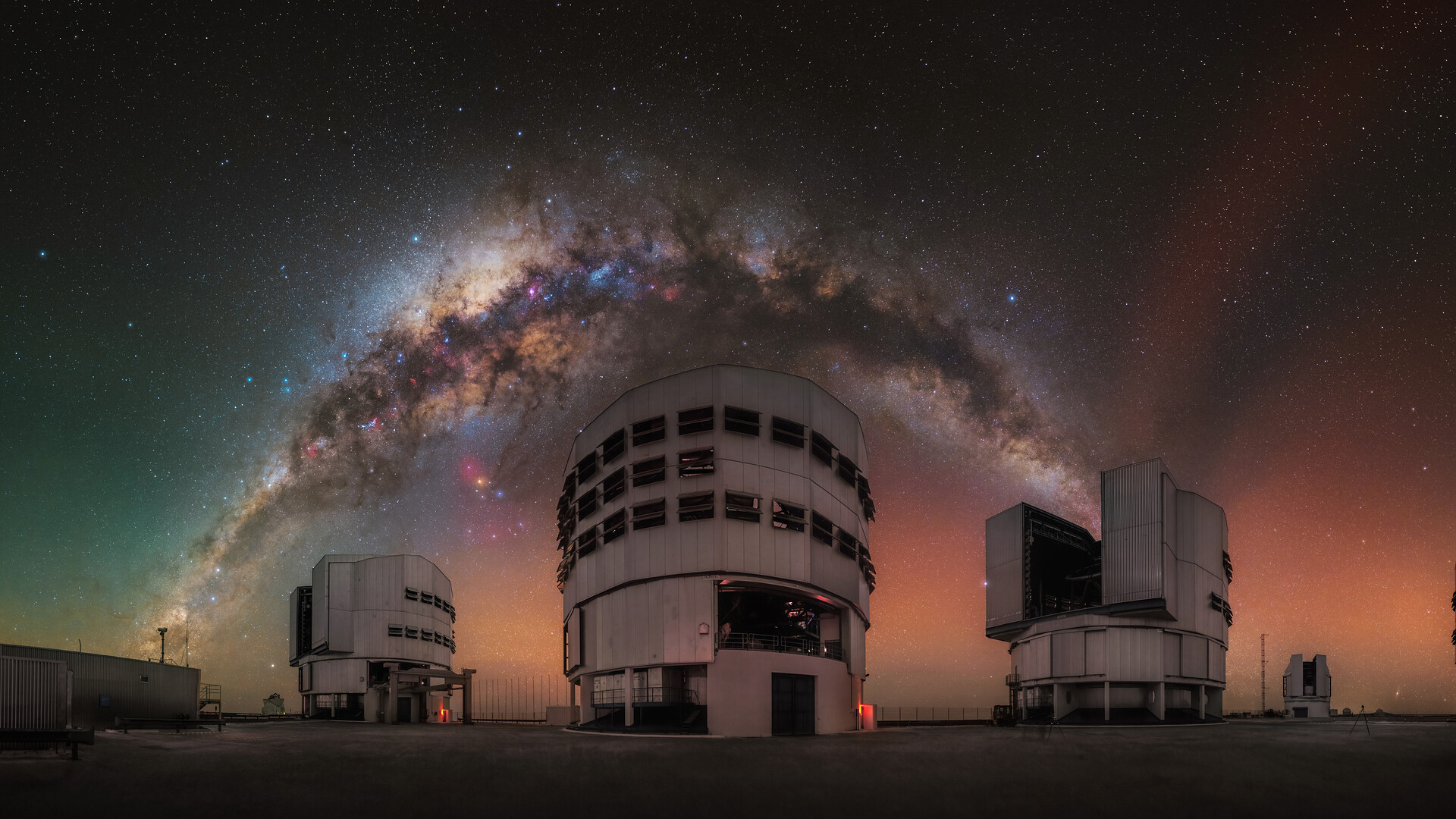 A glowing arch of the Milky Way galaxy can be seen in a red and orange and green night sky over three large towering buildings. 