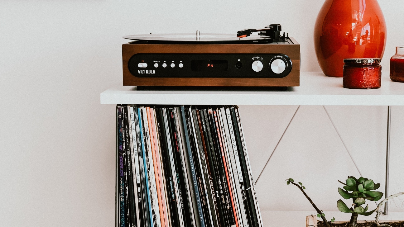 White wall with a brown record player sitting on a shelf with a stack of vinyls underneath