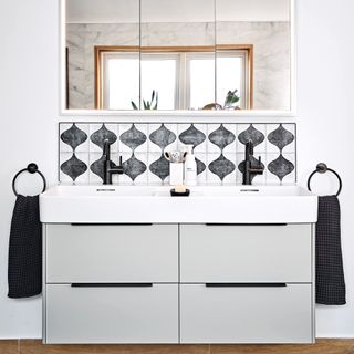 a bathroom with a twin basin vanity unit in light grey with a black and white patterned tile splashback and black taps, rectangular wall mirror, and black hand towels