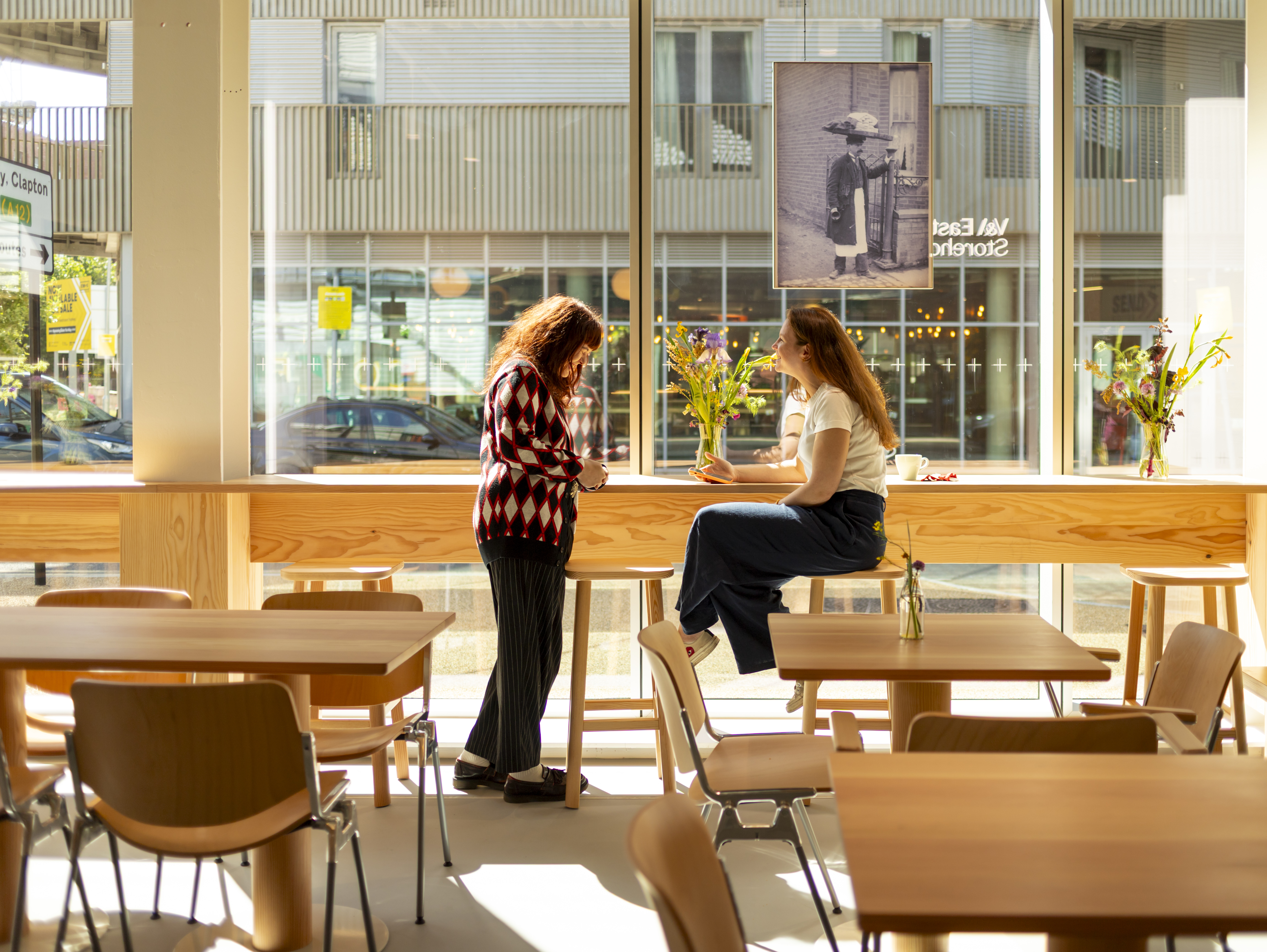 Two women talking in a light and airy cafe space with big windows and pale fir tables and chairs. There is a vase of fresh wildflowers between them
