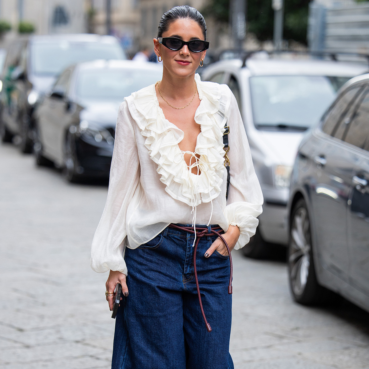 A woman wears a ruffled blouse and sunglasses with long denim shorts.