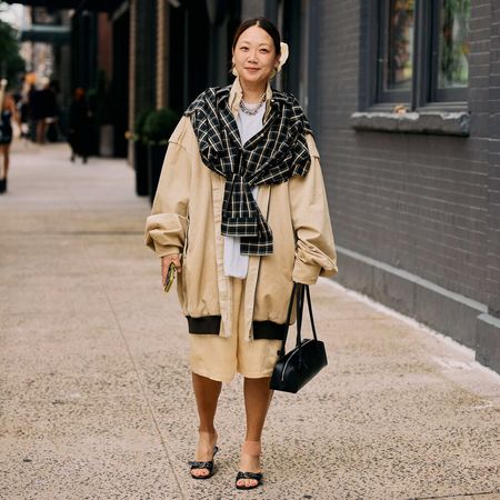 Woman wears oversized bomber, khaki trouser shorts, button down, plaid scarf, black handbag, and sandals.
