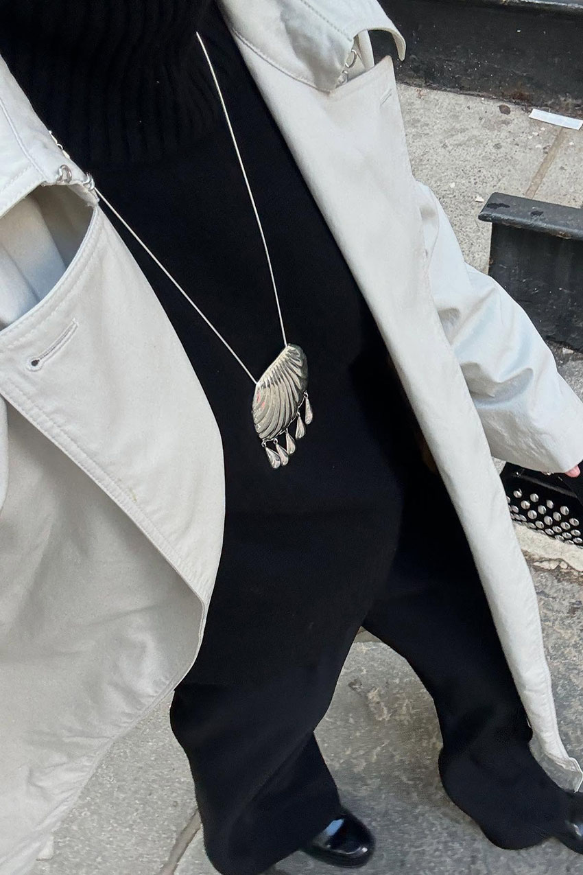 a woman wearing a khaki coat, black sweater, black pants, and silver pendant necklace