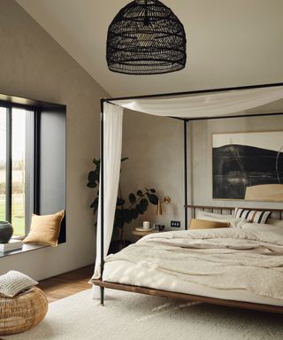 A neutral bedroom with a black four poster bed, a large window with a bench seat