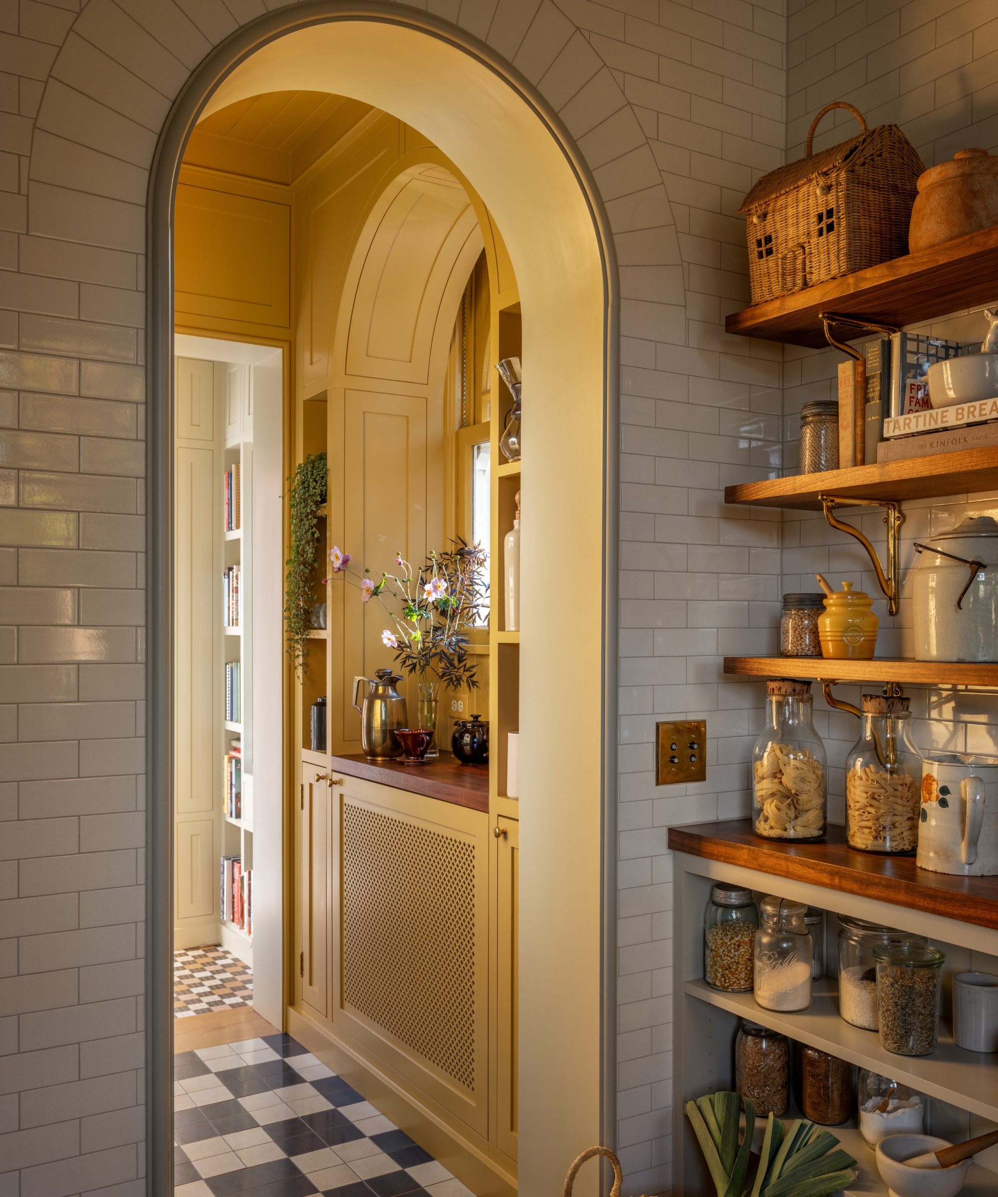 The corner of a small white kitchen with wooden open shelves serving as a pantry with an arched doorway looking into a yellow connecting room