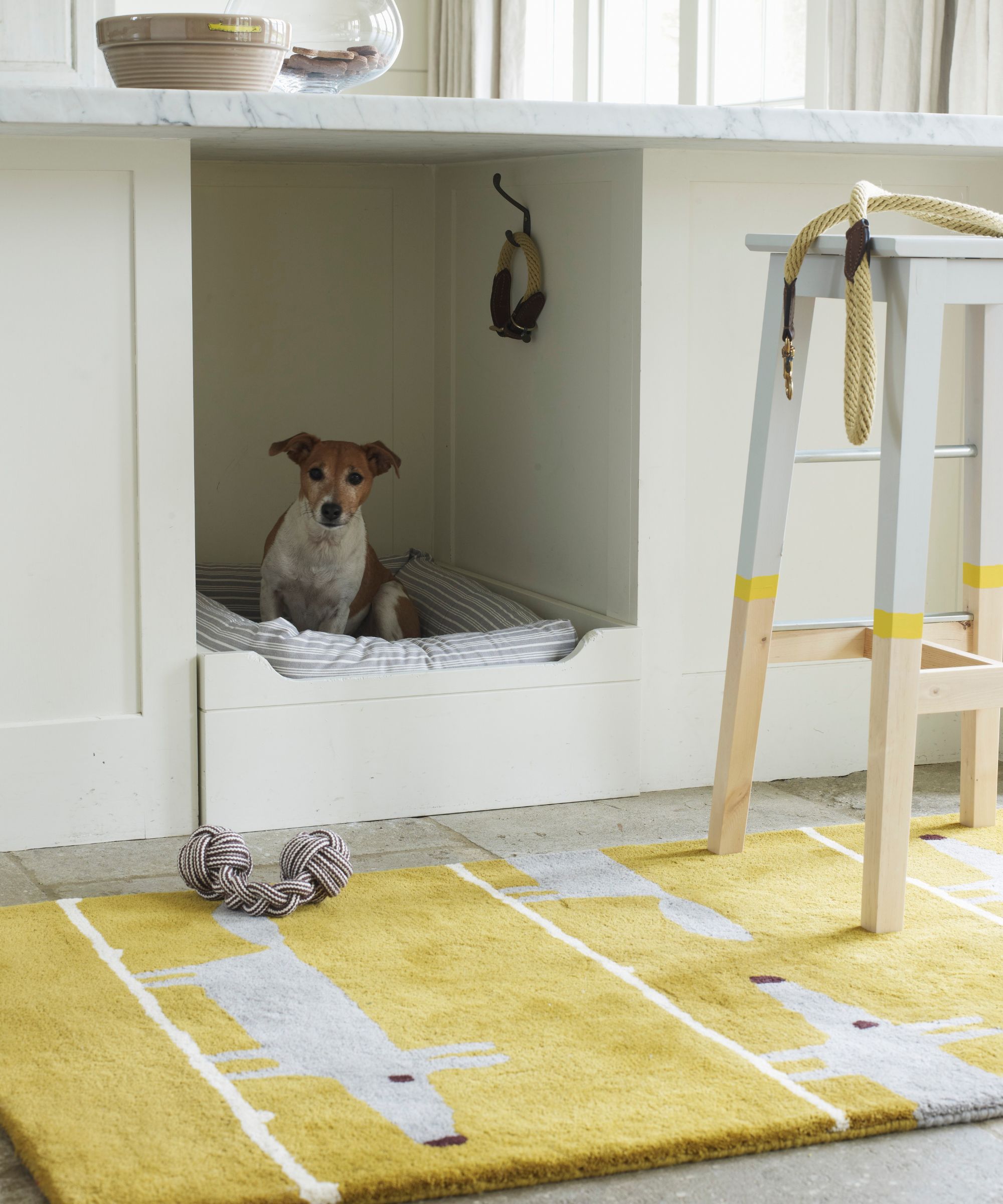 A small white and tan terrier dog, pictured in a built-in dog bed underneath a white kitchen island with marble countertop. On top of the countertop is a large ceramic bowl alongside a large glass bowl, and in the foreground is a bright yellow rug decorated with grey dogs, and a grey, yellow, and wood stool