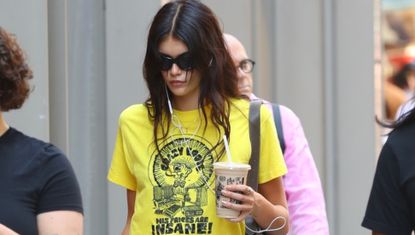 Kaia Gerber walks in New York City carrying a coffee while wearing the summer white skirt trend and black ballet flats