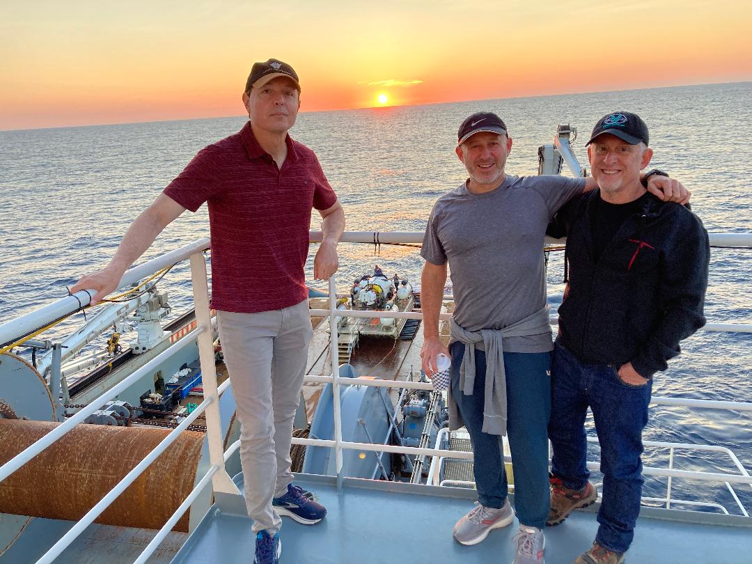 Crewmates Dylan Taylor, Randy Brunschwig and Alan Stern stand on the deck of a ship with the sun setting over the open ocean behind them.