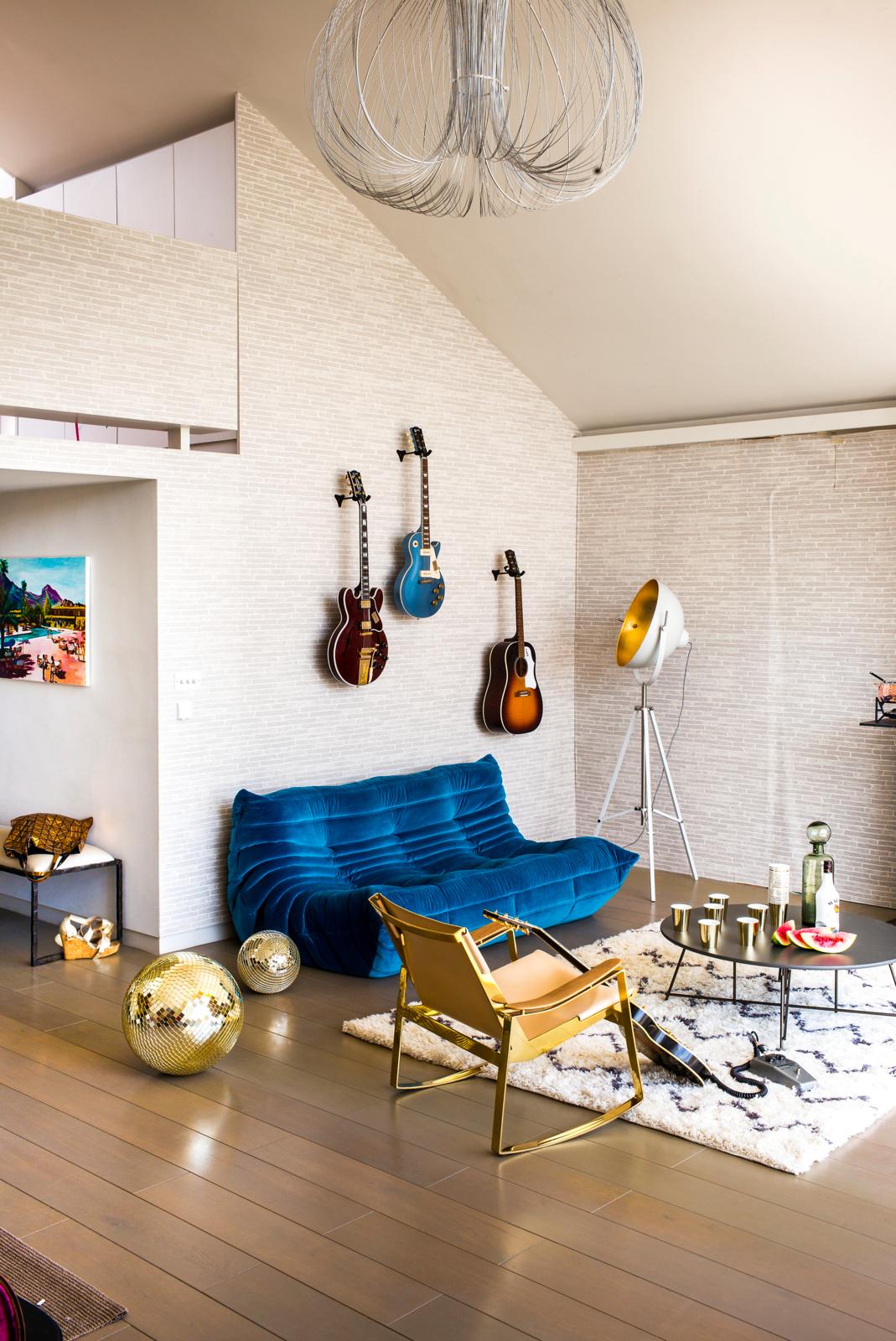 Image of an open living room in a beige colorway with a jewel-toned blue sofa and three guitars hanging on the wall. There are two gold disco balls on the ground and a gold accent chair.