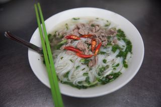 A large bowl of pho with noodles and strips of beef and red chiles on top with green chopsticks and a spoon on the side