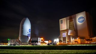 A white, rounded point cylyictrical capsule is rolled by a giant cube building.