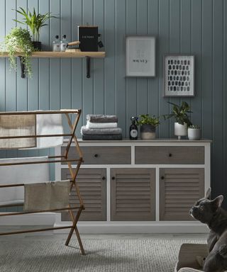 A nautically-inspired laundry room with teal-colored wall paneling, bamboo airer and sideboard with louvre doors, French bulldog in forefront