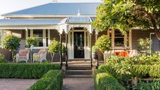 The facade of a home with lots of plants scattered around as well as a black traditional style door. There is patio furniture on the left and right side of the home. 