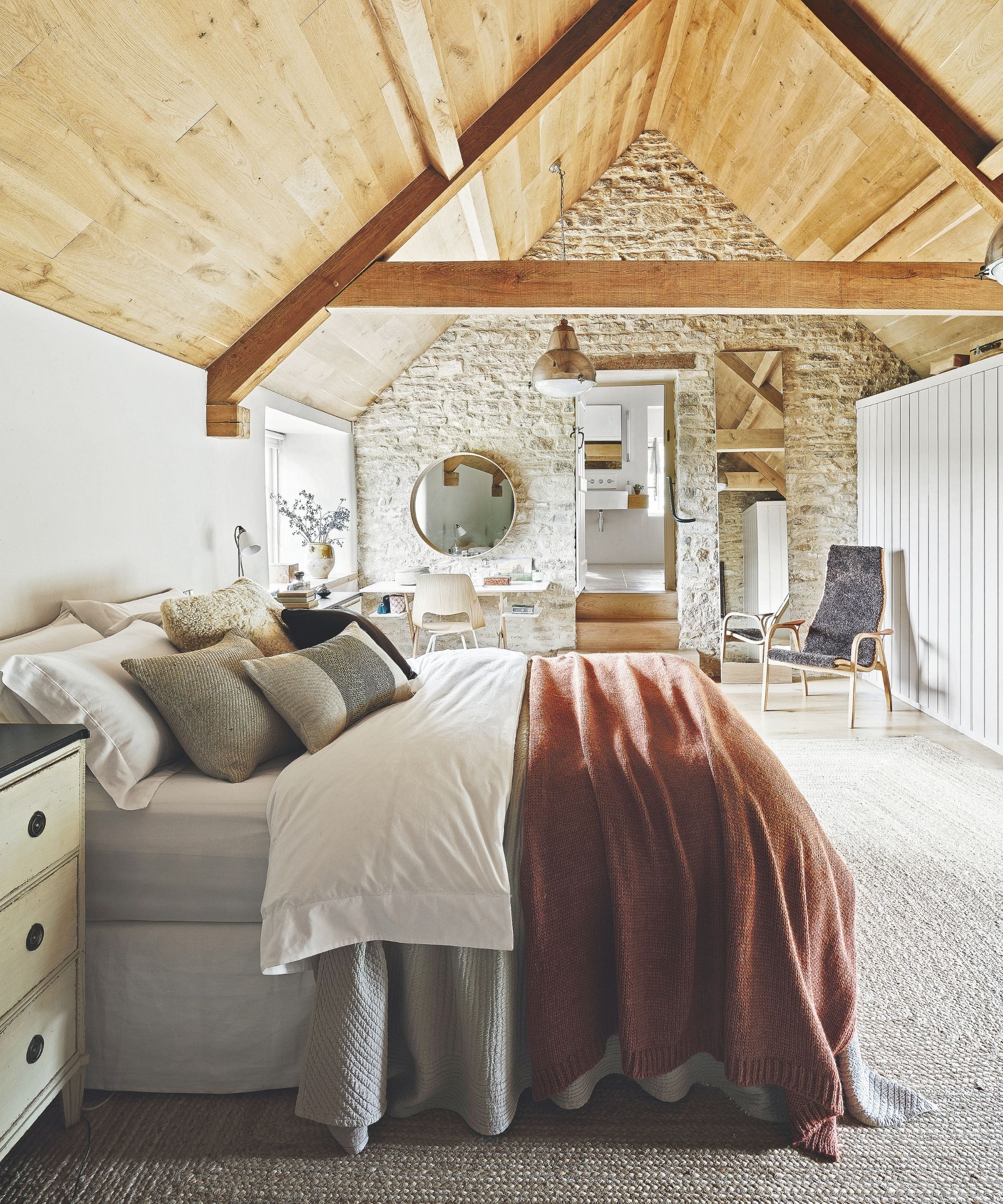 Attic bedroom with wood panelled ceiling, double bed with assorted blankets, bedding and cushions, exposed brick wall, pale grey carpet, and circular mirror above a white desk beside a window.