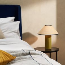 Bedside table lamp on round wooden table next to bed with golden pillow on top