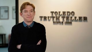 Ned (Domhnall Gleeson) poses, arms crossed, in front of the Toledo Truth Teller sign in a scene from Peacock's "The Paper"