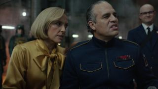 Toni Collette (lady on the left wearing a posh yellow blouse) and Mark Ruffalo (man on the right with slicked back hair and wearing a blue general-like uniform) in the movie Mickey 17.