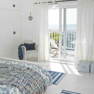 coastal bedroom with blue and white scheme, balcony view of beach, white painted rattan chair, chrome pendant light, blue and white striped rugs