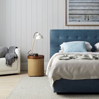 coastal bedroom ideas with blue upholstered bed, white tongue and groove walls, artwork above bed, blue and stone bedding, round leather and wood side table, white and wood lamp, white armchair, wooden floor