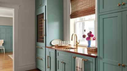 Kitchen with cabinetry painted in Benjamin Moore's Stratton Blue, a wooden countertop with a gold faucet, a rattan blind on top of the window, and a vase of pink flowers on the windowsill.