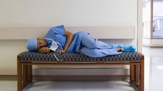 A nurse wearing blue scrubs asleep on her side on a bench in a hospital