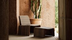 Image of a dark patio corner with pink plaster walls and a large terracotta pot that has a cactus in it. There is a plastic, basketweave outdoor armchair and matching ottoman.