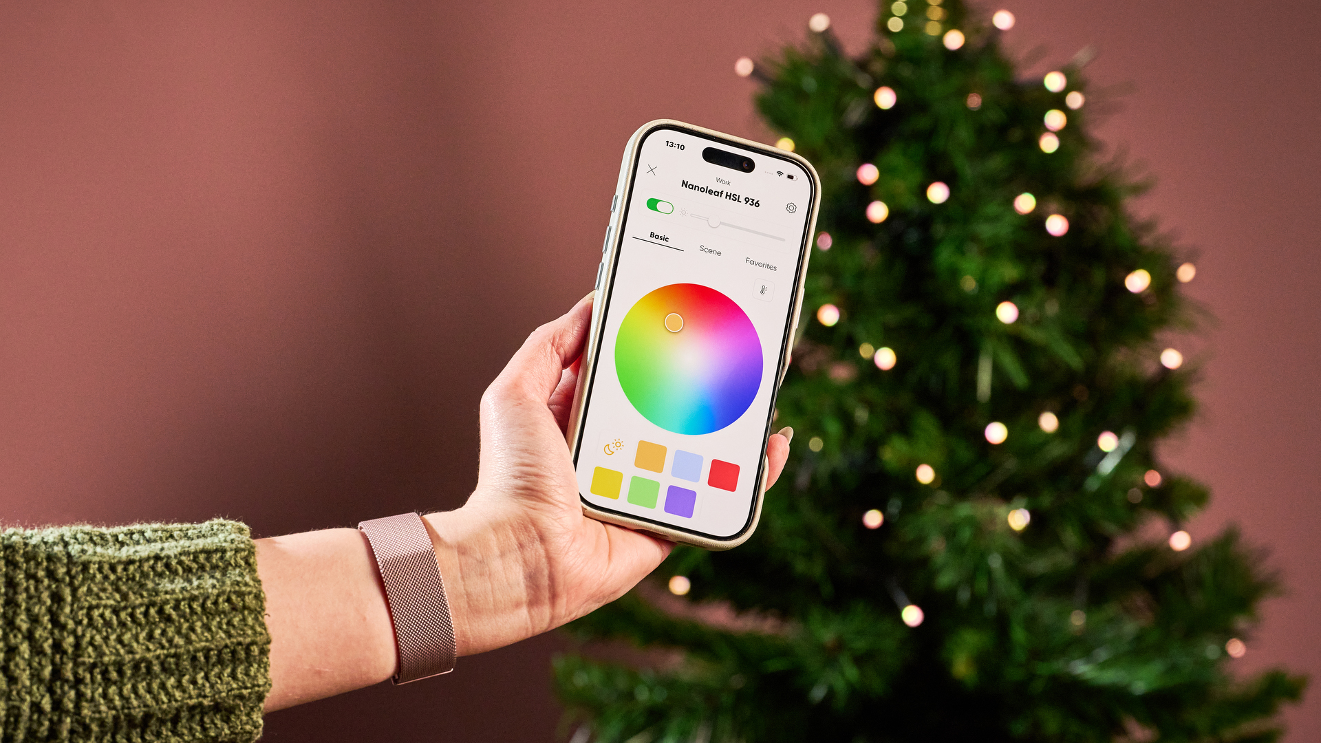 A woman holds a smartphone showing the Nanoleaf app. In the background, the Nanoleaf Smart Holiday String Lights are glowing on a Christmas tree.