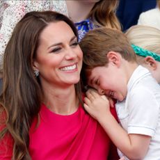 Princess Kate wearing a pink dress laughing as Prince Louis puts his head on her shoulder 