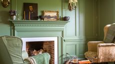 Living room with fireplace and walls painted in Calke Green by Farrow & Ball, two armchairs, and a mantelpiece with antique paintings, vases and candlesticks